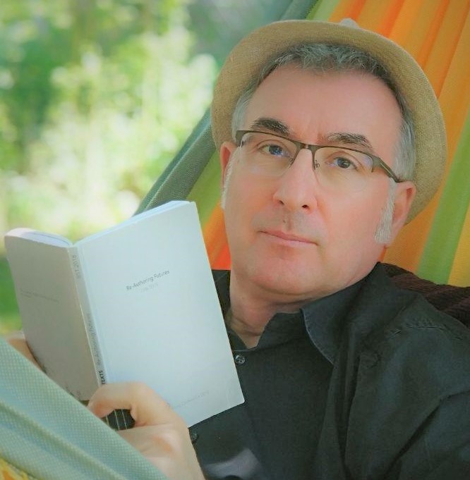 Ένα άτομο που φοράει γυαλιά και καπέλο κάθεται σε μια αιώρα, κρατώντας ένα βιβλίο με τίτλο «Re Authoring Futures». Το φόντο έχει απαλά, θολά χρώματα που υποδηλώνουν ένα φυσικό περιβάλλον.