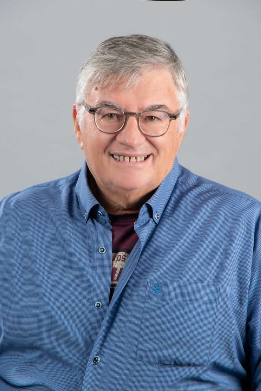 Ένα χαμογελαστό άτομο με γυαλιά και γκρίζα μαλλιά, που φοράει μπλε πουκάμισο με κουμπιά, σε φόντο ανοιχτό γκρι χρώμα.