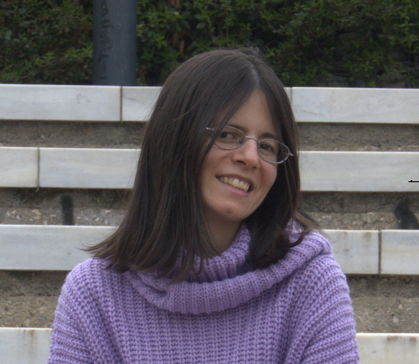 Ένα χαμογελαστό νεαρό άτομο με μακριά σκούρα μαλλιά και γυαλιά κάθεται σε πέτρινα σκαλοπάτια, φορώντας ένα μοβ πουλόβερ.